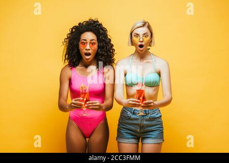 excited interracial women in summer outfit holding cocktail glasses and looking at camera on yellow Stock Photo