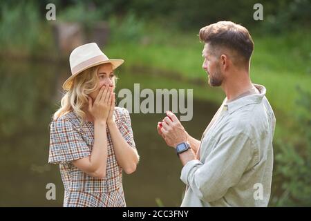 Waist up portrait of man proposing to beautiful girlfriend while standing by lake outdoors in sunlight, copy space Stock Photo