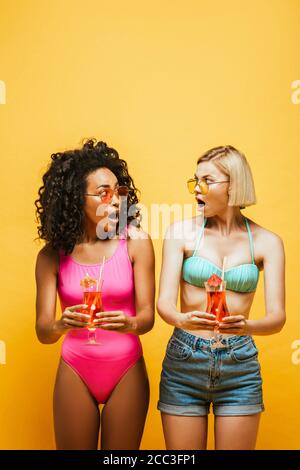 shocked interracial friends in summer outfit holding cocktails and looking at each other on yellow Stock Photo