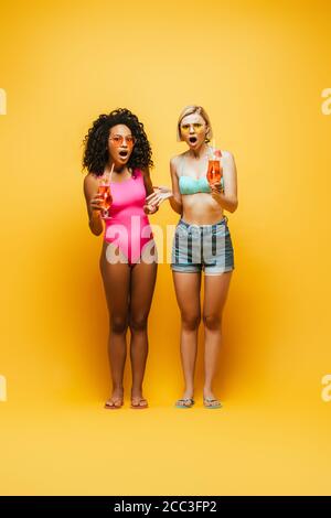 full length view of young interracial women in summer outfit holding cocktail glasses and looking at camera on yellow Stock Photo