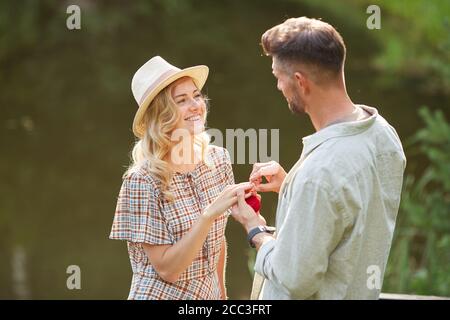 Waist up portrait of beautiful young woman accepting marriage proposal while standing by lake in sunlight, copy space Stock Photo