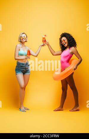 excited interracial women in summer outfit clinking cocktail glasses and looking at camera on yellow Stock Photo