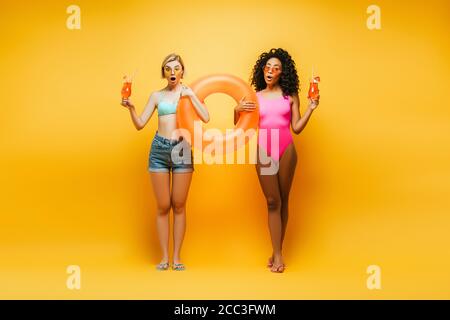 surprised blonde woman in shorts with african american friend in swimsuit holding swim ring and cocktail glasses on yellow Stock Photo