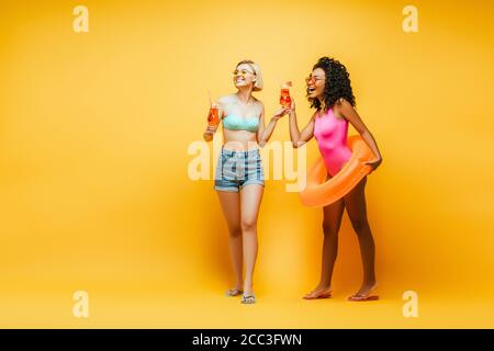 blonde woman with african american friend holding cocktail glasses and swim ring laughing while looking away on yellow Stock Photo