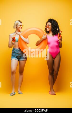 young interracial women in summer outfit holding swim ring and cocktail glasses while looking at each other on yellow Stock Photo