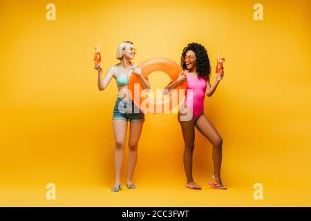 young interracial women in summer outfit posing with swim ring and cocktail glasses while looking at each other on yellow Stock Photo