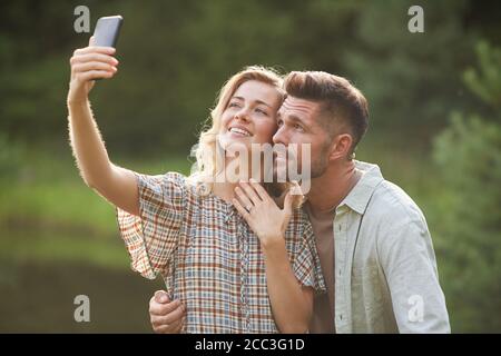 Waist up portrait of beautiful adult couple taking selfie with engagement ring after marriage proposal during romantic date outdoors Stock Photo