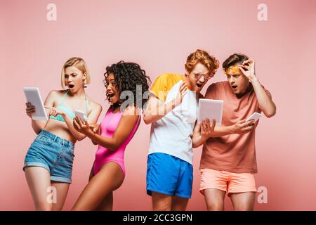 shocked multicultural friends in summer outfit using digital devices isolated on pink Stock Photo