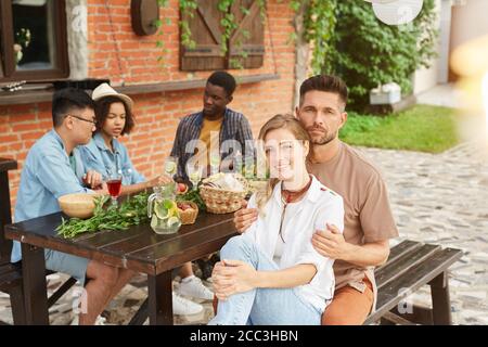 Multi-ethnic group of beautiful young people enjoying dinner outdoors in sunlight, focus on smiling couple in foreground, copy space Stock Photo