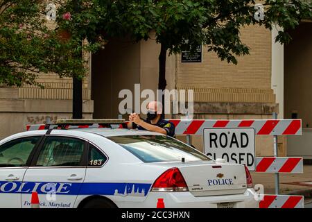 Frederick, MD, USA 08/14/2020: A young caucasian male police officer is wearing a face mask while on duty. He is standing by his police car in front o Stock Photo