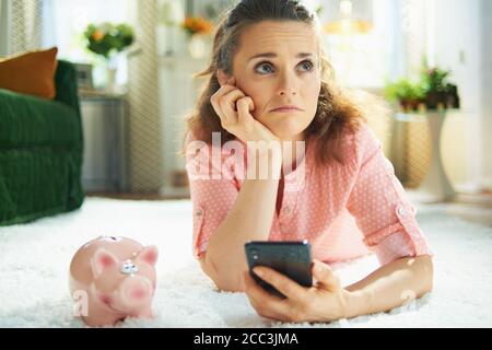 sad modern 40 years old woman in blouse and white pants with piggy bank searching for carpet cleaning service on a smartphone while laying on white ca Stock Photo