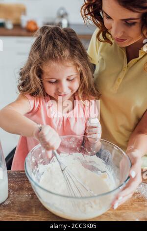 high angle view of mother touching glass bowl while daughter kneading dough with whisk Stock Photo
