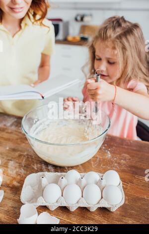 cropped view of mother reading cookbook while daughter kneading dough with whisk in glass bowl, selective focus Stock Photo