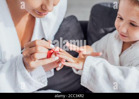 cropped view of woman applying red enamel on fingernails of child in white bathrobe Stock Photo