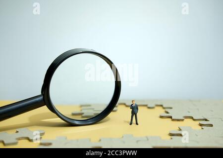 Business strategy conceptual photo - Miniature businessman standing behind magnifier glass in the middle of jigsaw puzzle Stock Photo