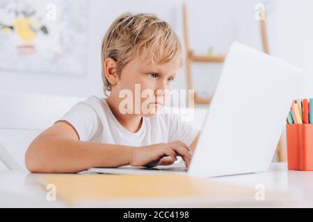 selective focus of concentrated boy in white t-shirt sitting at table and using laptop Stock Photo