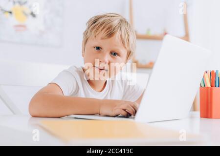 selective focus of kid in white t-shirt looking at camera while sitting at table and using laptop Stock Photo