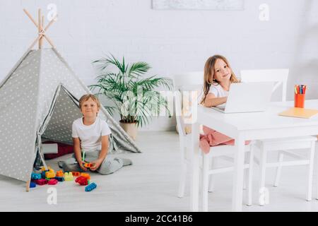 girl using laptop at table near brother sitting near kids wigwam and building blocks Stock Photo