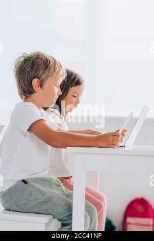 side view of brother and sister in pajamas sitting at while table and using laptops together Stock Photo