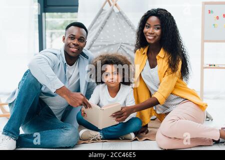 african american parents and curly kid holding book while sitting on floor Stock Photo
