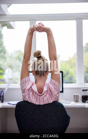 Rear View Of Woman Working From Home On Computer  In Home Office Stretching At Desk Stock Photo
