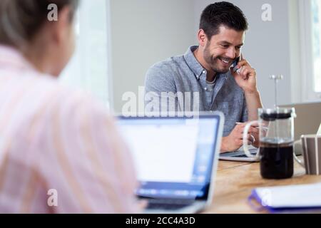 Business Couple Working From Home Sitting At Table During Pandemic Lockdown Using Mobile Phone Stock Photo