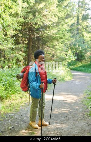 Portrait of African woman with backpack and sticks smiling at camera while standing in the forest Stock Photo