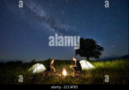 Beautiful view of night starry sky over meadow with hikers near illuminated camp tents. Tourists sitting near campfire under magical blue sky with stars. Concept of travelling, hiking and camping. Stock Photo