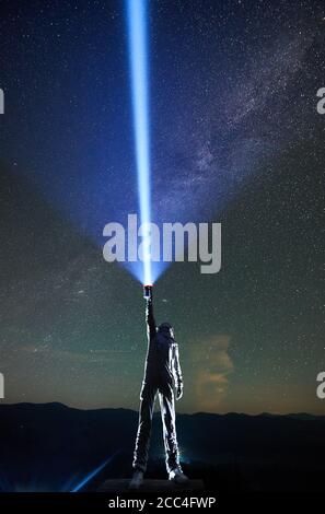 Space traveler in space suit and helmet illuminating beautiful starry sky with flashlight while standing under magical night sky with Milky Way. Concept of light, astronomy, cosmonautics Stock Photo