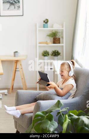 Girl with down syndrome sitting on armchair in headphones watching something on tablet pc and laughing Stock Photo