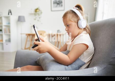 Down syndrome girl in wireless headphones using digital tablet while resting on armchair in the living room Stock Photo