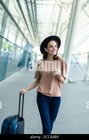 Young woman carry baggage speak via airpods with phone in hands walking to airport terminal Stock Photo