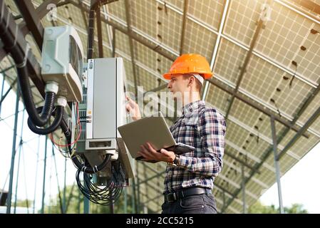 Expert is inspecting quality of solar panels. Man in hardhat standing with laptop near electrical equipment. Solar station sunny power electricity. Innovation future. Stock Photo