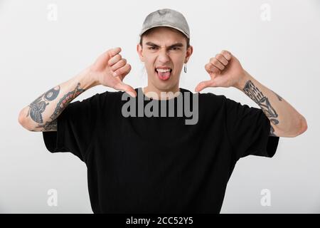 Portrait of irritated young man wearing black clothes showing thumb down and sticking out his tongue over white background Stock Photo