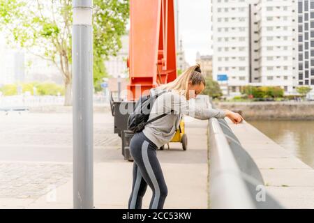 An athletic woman in a grey sweater resting and leaning on the bridge railing with headphones around her neck. Stock Photo