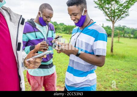 Group of cheerful African friends with facemasks having a drink and using their phones in a park Stock Photo