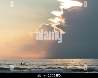 surfer waiting for waves in a overcast evening, the sun's rays filter through the clouds Stock Photo