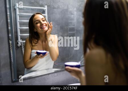 Smiling young woman applying cream to face and looking to mirror at home bathroom Stock Photo
