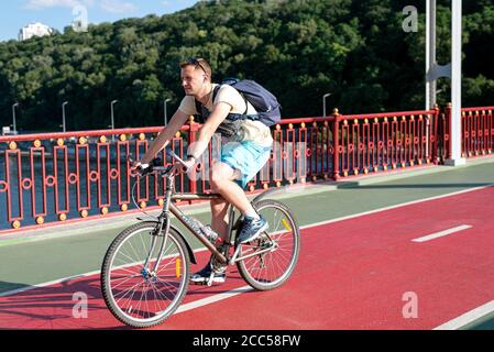 Kyiv, Ukraine - 11 July, 2020. Pedestrian bridge. Young man riding bicycle to work enjoying music using earphones on city bridge Stock Photo