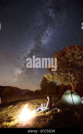Young happy woman traveller sitting at campfire beside camping and illuminated tourist tent under night sky full of stars and Milky way. On the background starry sky, mountains and big tree Stock Photo
