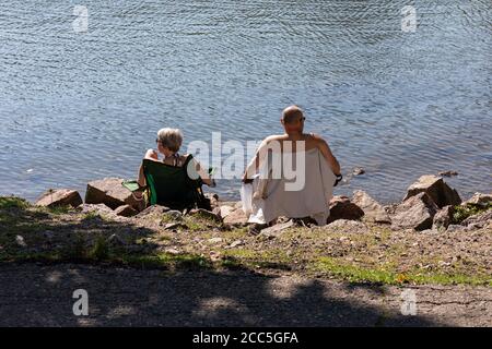 Middle-aged couple sitting on portable camping chairs by water Stock Photo