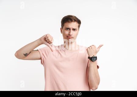 Image of dissatisfied caucasian man in basic t-shirt showing thumb down and pointing finger aside isolated over white background Stock Photo