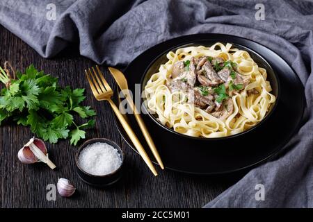 beef stroganoff cooked with sour cream mushrooms sauce, served with egg noodles in a black bowl with golden fork and knife on a dark wooden table, clo Stock Photo