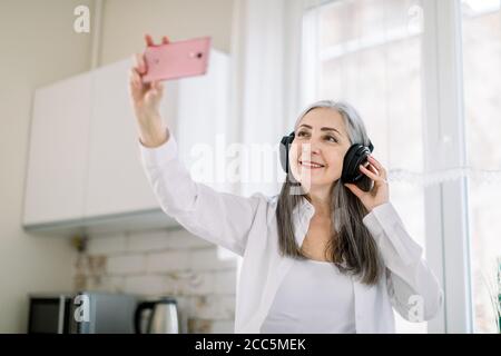 Beautiful retired gray haired woman in white shirt standing in the kitchen, listening to music in headphones and using media applications on her Stock Photo