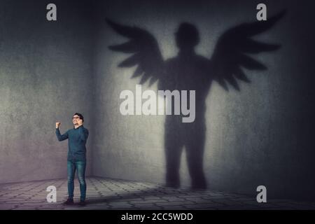 Successful businessman, raising hands up, celebrating victory, casting a superhero shadow with angel wings on a dark room wall. Self overcome, leaders Stock Photo