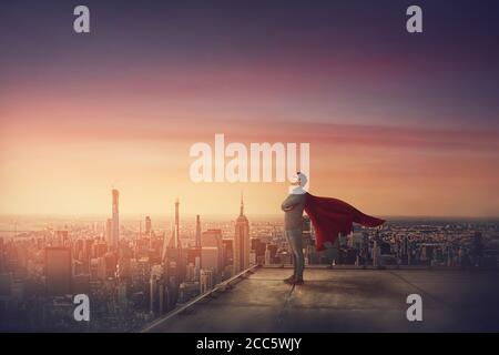 Conceptual sunset scene, business person with red cape stands confident on rooftop looking determined as a superhero over city horizon. Ambition and l Stock Photo