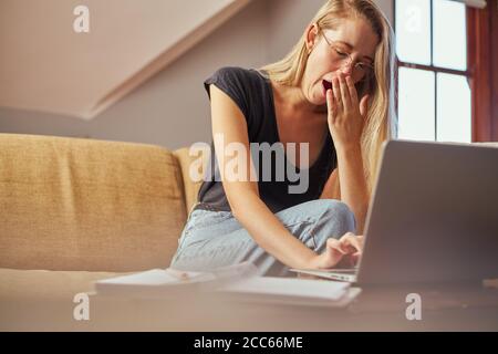 Yawning young woman working from home on sofa in living room Stock Photo