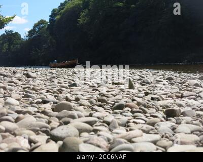 Foreground of a rocky shore looking out to a small boat on the River Tay; Dunkeld, Perthshire, August 2020 Stock Photo