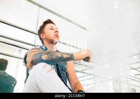 Portrait of modern tattooed art dancer teenager dressed in jeans vest smokes an Electronic cigarette in mirror dance hall Stock Photo