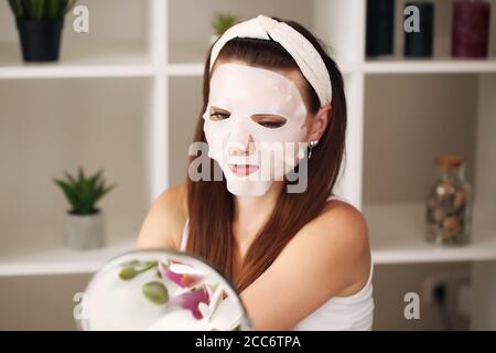 Beautiful young woman putting face moisturizing mask in front of a mirror and looking at her own reflection. Close up Stock Photo